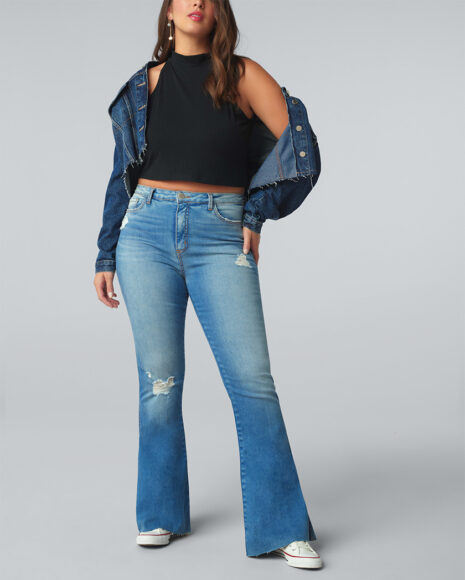 Uma mulher usando cropped preto gola alta, com uma jaqueta jeans escura caída nos ombros, calça jeans reta e tênis all star branco