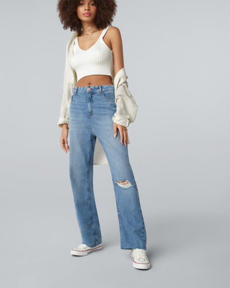 Uma mulher vestindo um cropped de lã branco com um casaco por cima, uma calça jeans reta clara com rasgos na perna e um all star branco 