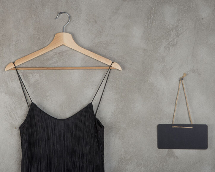 Um vestido preto de festa pendurado em um cabide em um fundo ao lado tem uma bolsa preta com a alça dourada em um fundo cinza 