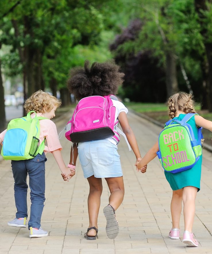 três crianças de mãos dadas andando até a escola e usando mochila escolar